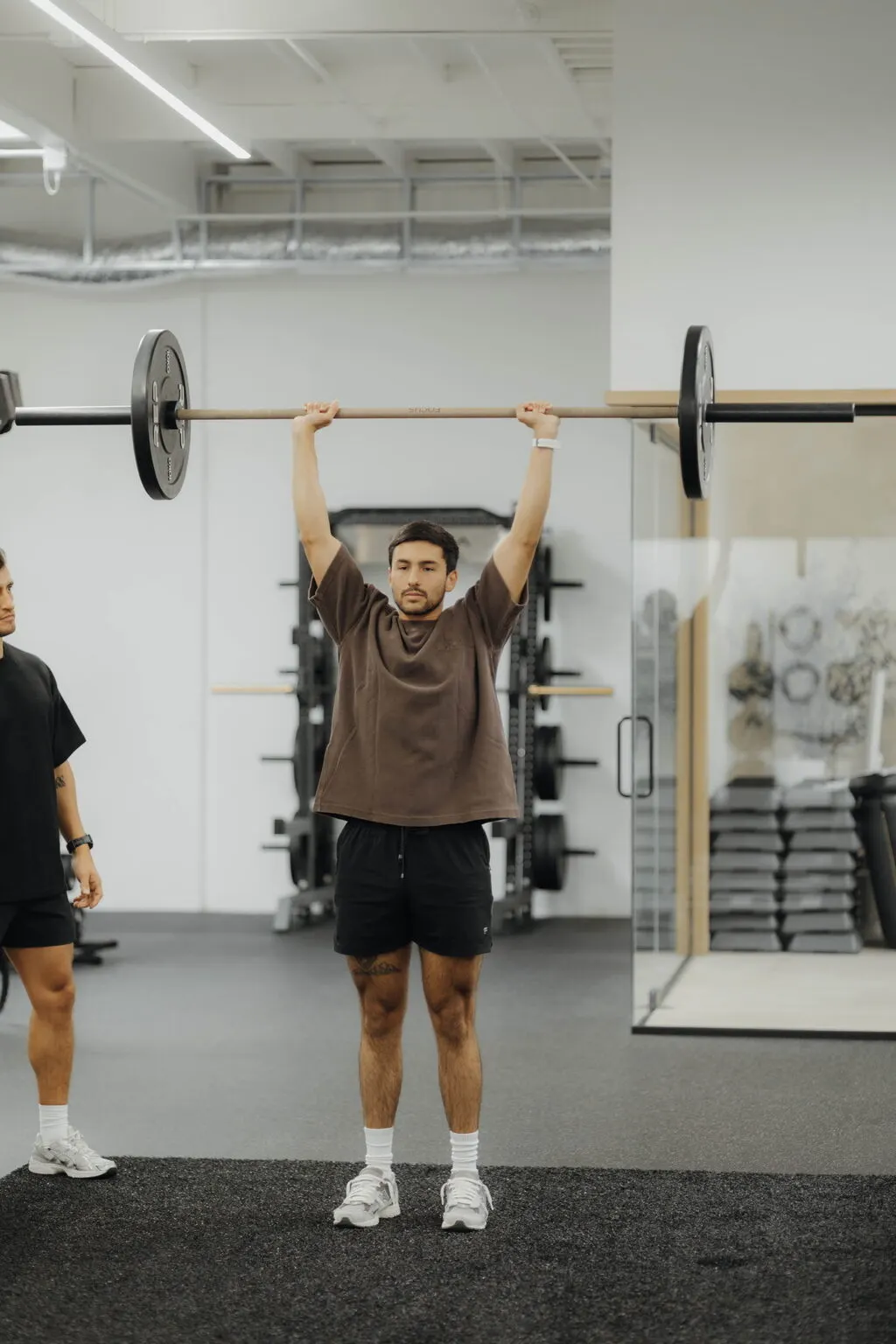 A client pressing a barbell overhead while Coach Santi watches his technique, 1-on-1 strength training at Focus Athletics in Costa Mesa